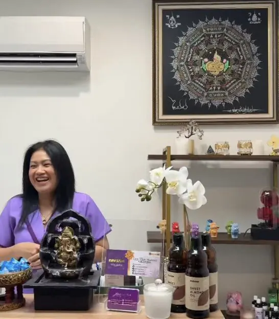 Suw smiling at the Lux Thai reception desk, with orchids and a framed Thai artwork behind